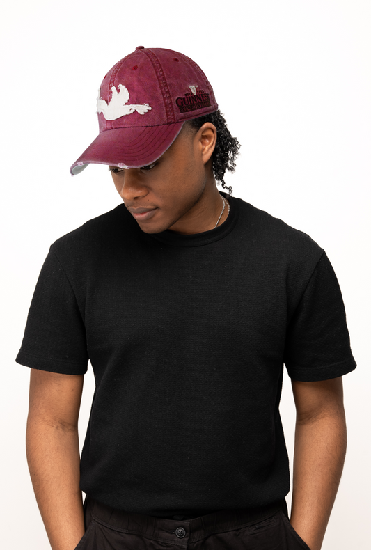 A person in a Guinness Webstore US Guinness Red Toucan Cap, black t-shirt, and black pants stands against a white background, looking down. The cap’s subtle distressed style adds a vintage touch to the overall look.