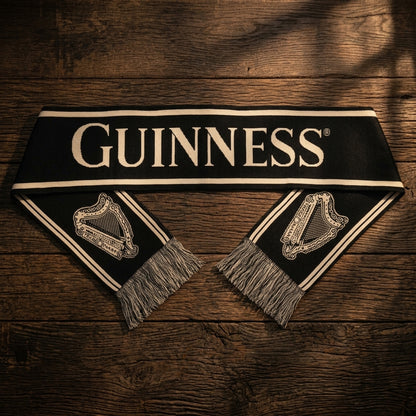 The Guinness Black & Gold Sports Scarf – Vintage Harp Knit by Guinness, featuring a vintage harp design and fringe ends, is laid flat on a wooden surface.