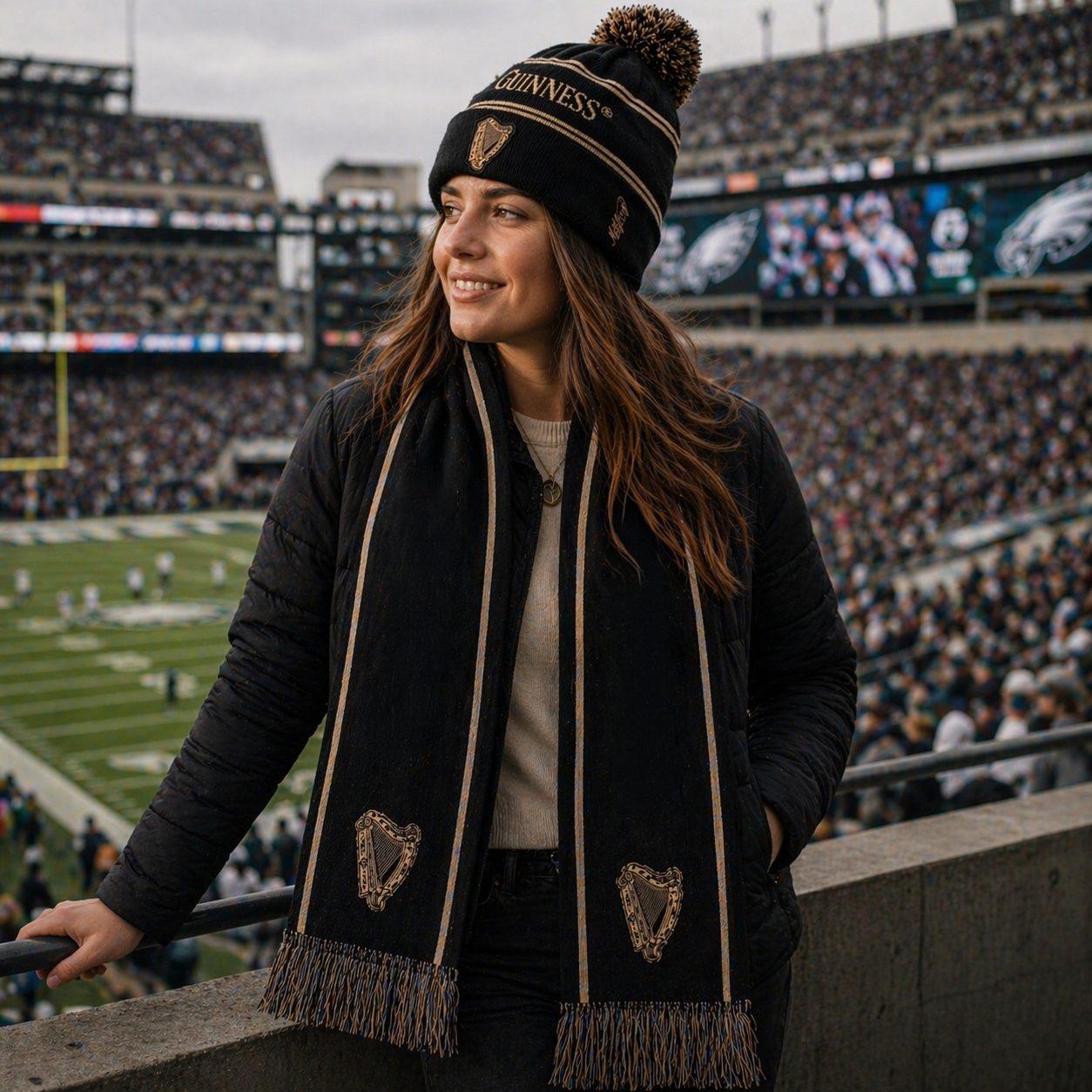 A woman wearing the Guinness Black & Gold Sports Beanie – Vintage Harp Knit stands in a stadium, watching a football game as the crowd buzzes behind her.