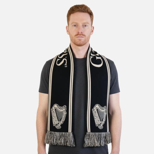 A man with short red hair wears a black t-shirt and the Guinness Black & Gold Sports Scarf – Vintage Harp Knit, featuring fringed ends.