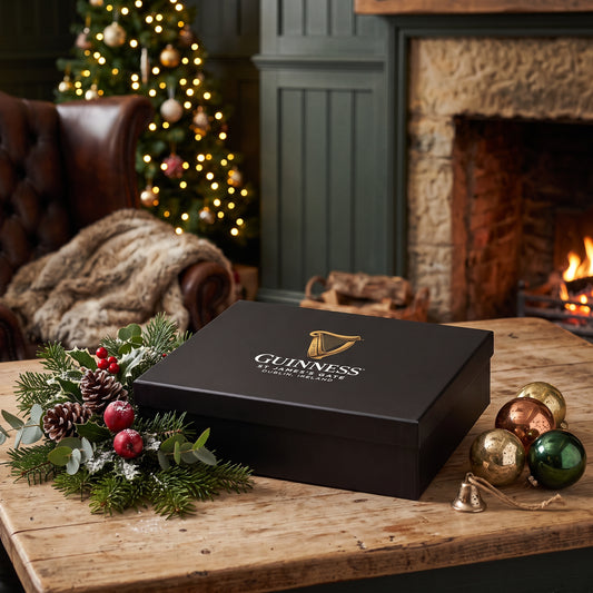A black rectangular Guinness Gift Box - Medium with a gold harp logo sits on a wooden table, decorated with holiday greenery and ornaments in front of a fireplace and Christmas tree.