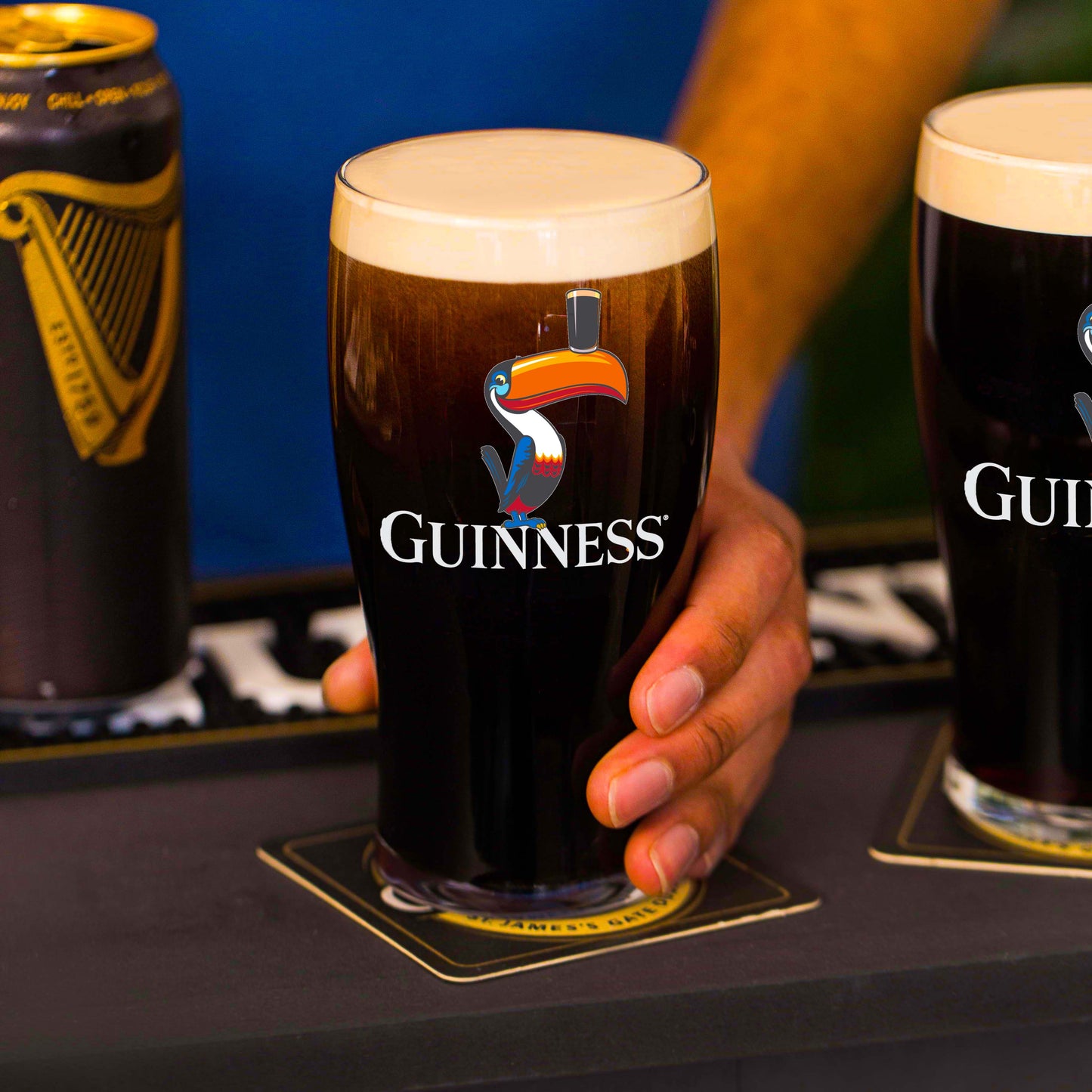 Someone holds a Guinness Toucan Pint Glass on a coaster, with a Guinness can and another pint glass—possibly also from Guinness—partially visible nearby.
