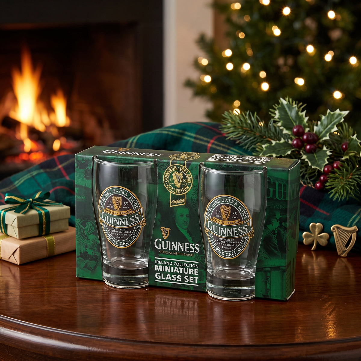 The Guinness Ireland Collection Mini Glass Set from Guinness Webstore US is shown in a green box on a festive table with holiday decor, a plaid blanket, and a cozy fireplace.