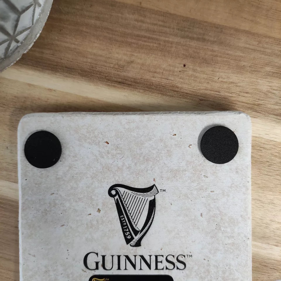 A Guinness Gilroy Toucan Stone Coaster with two black rubber feet sits on a wooden surface beside the base of a glass object.