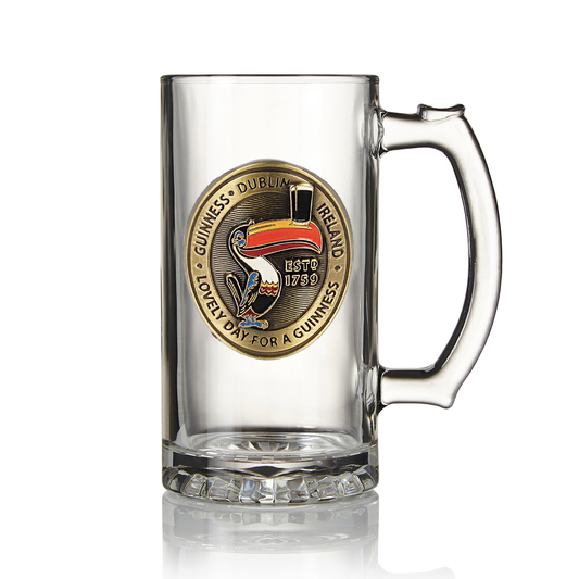 A clear glass Guinness Gilroy Toucan Tankard with a Guinness Dublin Ireland badge featuring the iconic John Gilroy design of a toucan and beer pint. Text on the badge reads: "Established 1759" and "Lovely day for a Guinness." A perfect addition to any Guinness collectible enthusiast's collection, available from Guinness Webstore US.