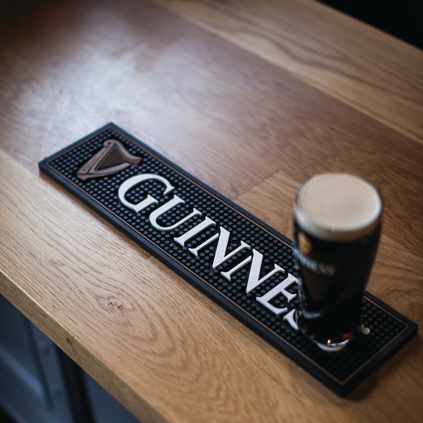 A pint of Guinness sits on the Guinness Harp Bar Mat by Guinness, perfectly placed on a wooden bar—an ideal accent for your home bar accessories.