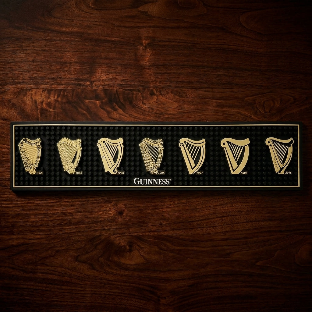 The Guinness Evolution of the Harp PVC Bar Mat features seven distinct gold harp logos—from 1862 to 2016—displayed on a wooden-look background, celebrating the iconic evolution of the Guinness brand and the logo through the years.