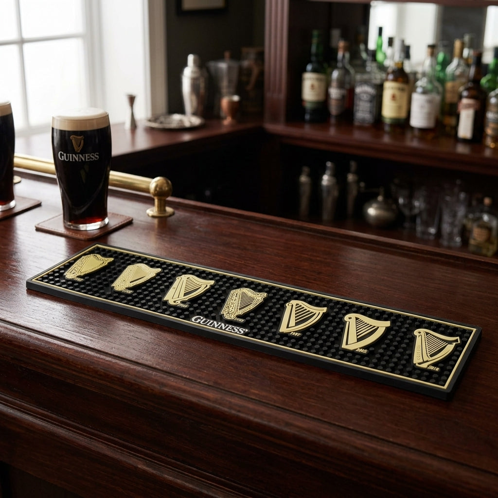 A wooden bar counter holds two pints of Guinness, with liquor bottles on shelves behind and a Guinness Evolution of the Harp PVC Bar Mat featuring seven gold harp Guinness logos with years from 1862 to 2016 and a central Guinness logo.