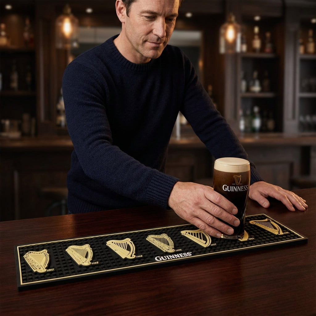 A man sets a pint of Guinness on the Guinness Evolution of the Harp PVC Bar Mat, features seven gold harp Guinness logos with years from 1862 to 2016 and a central Guinness logo, in a dimly lit pub.