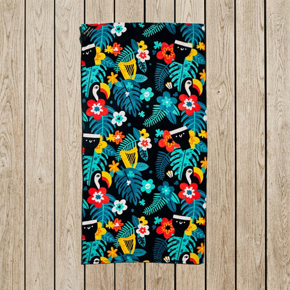 Guinness x Fatti Burke Hawaiian Beach Towel from Guinness Webstore US, featuring a colorful tropical pattern with toucans, leaves, and flowers, laid flat on a wooden floor.