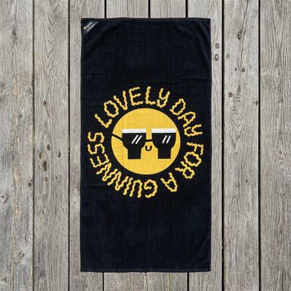 The Guinness x Fatti Burke Sun Beach Towel from Guinness Webstore US features a yellow smiley face with sunglasses and the words "LOVELY DAY FOR A GUINNESS" in a circular pattern on a black background, displayed on a wooden surface.