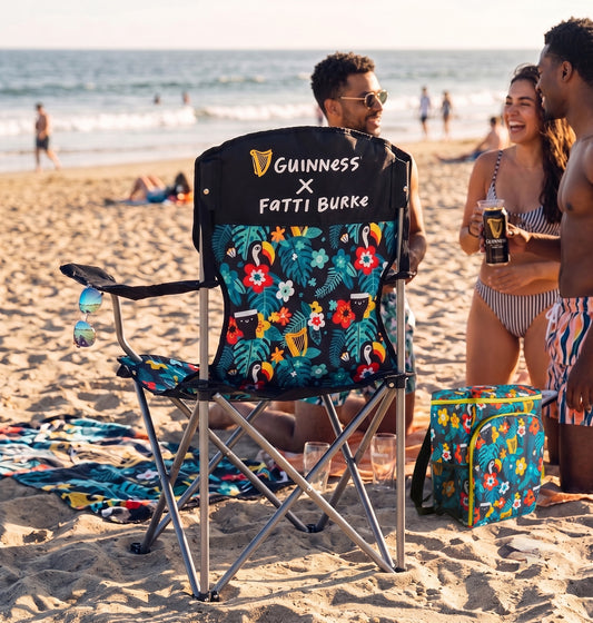 A vibrant Guinness x Fatti Burke Hawaiian Camping Chair from the Guinness Webstore US and a matching cooler are set on the sand near three people talking, with the ocean in the background.