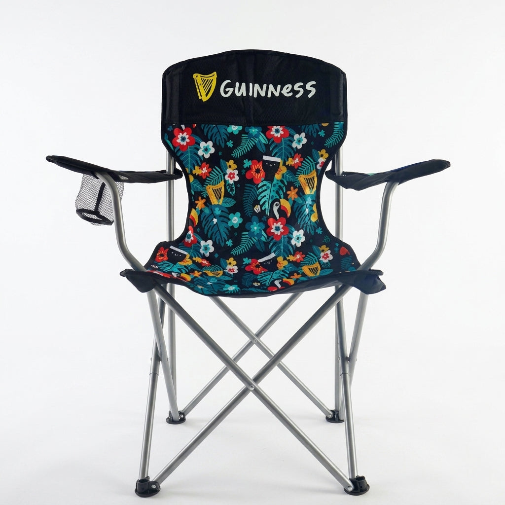 The Guinness x Fatti Burke Hawaiian Camping Chair from Guinness Webstore US showcases distinctive “Guinness” branding on a durable black frame with a vibrant tropical floral print fabric. Designed with comfortable armrests and a handy mesh cup holder on the left side, this chair is an ideal beach or outdoor companion. Photographed against a plain white background.
