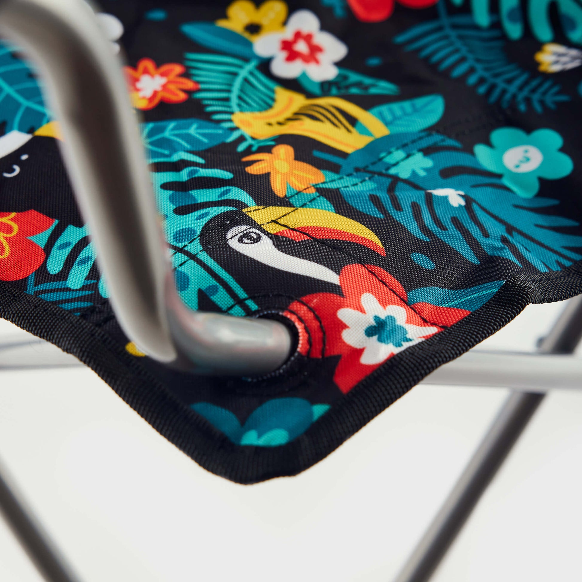 Close-up of the Guinness x Fatti Burke Hawaiian Camping Chair by Guinness, featuring a vibrant tropical print with leaves, flowers, and a toucan on a silver metal frame—ideal for pairing with your beach towel for summer fun.