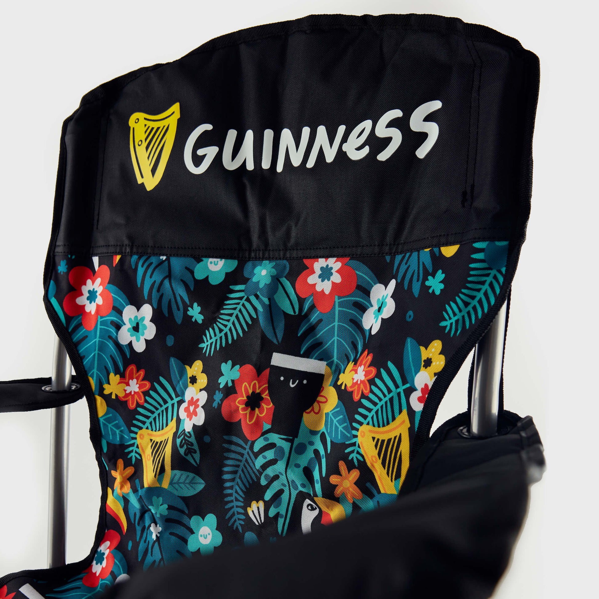 A close-up of the Guinness x Fatti Burke Hawaiian Camping Chair shows the Guinness logo on the black headrest and a vibrant floral backrest with toucan and pint glass illustrations, capturing the brand's iconic beach style.