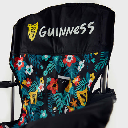 A close-up of the Guinness x Fatti Burke Hawaiian Camping Chair shows the Guinness logo on the black headrest and a vibrant floral backrest with toucan and pint glass illustrations, capturing the brand's iconic beach style.