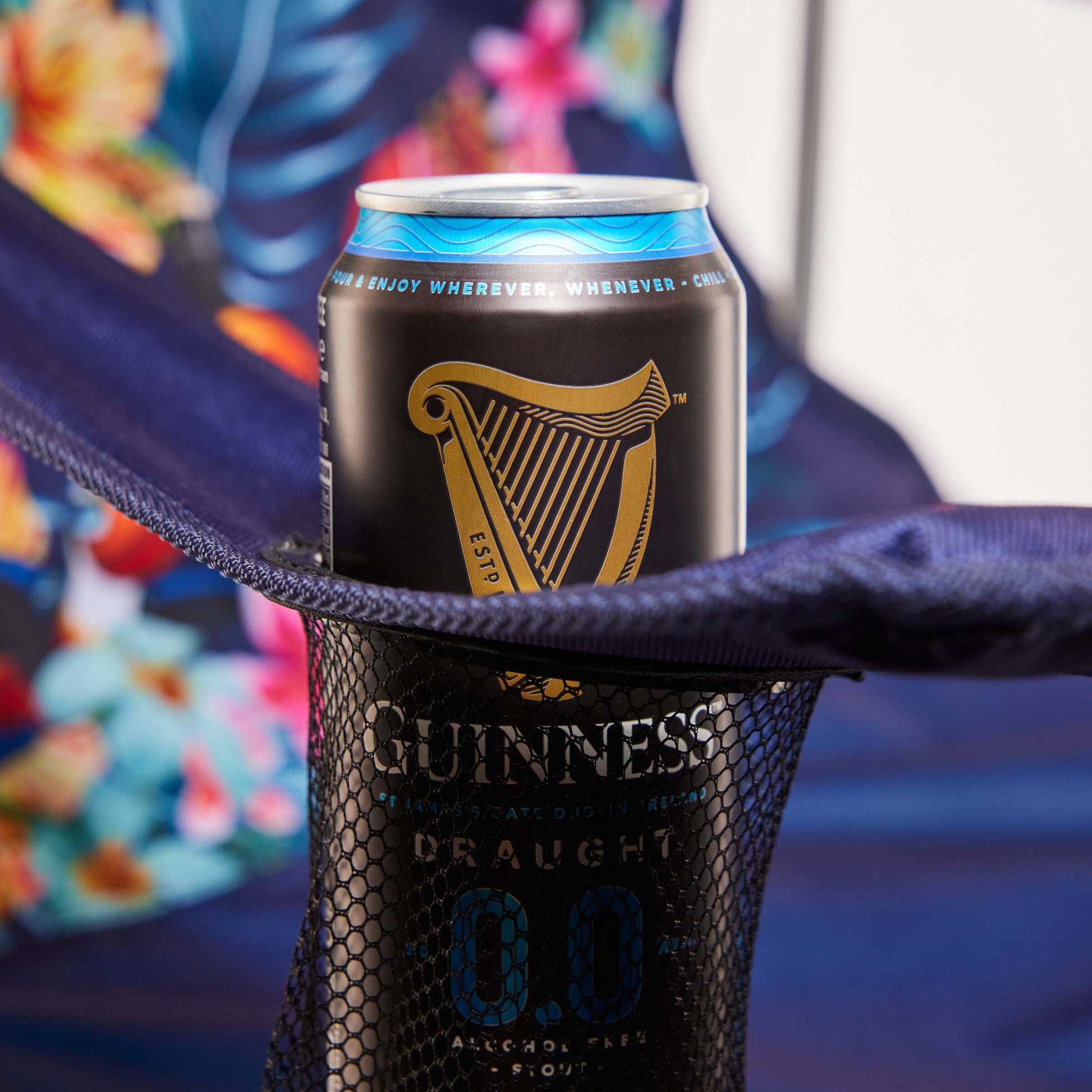 A can of Guinness 0.0 non-alcoholic beer rests in a mesh cup holder on the Guinness Toucan Hawaiian Beach Chair by Guinness Webstore US, with its vibrant floral pattern making it the perfect beach essential for your day out.