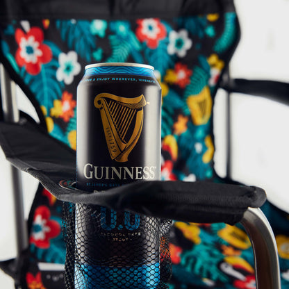A can of Guinness beer rests in the cup holder of a Guinness x Fatti Burke Hawaiian Camping Chair, featuring a colorful floral and leaf pattern, next to a vibrant beach towel draped over the armrest.