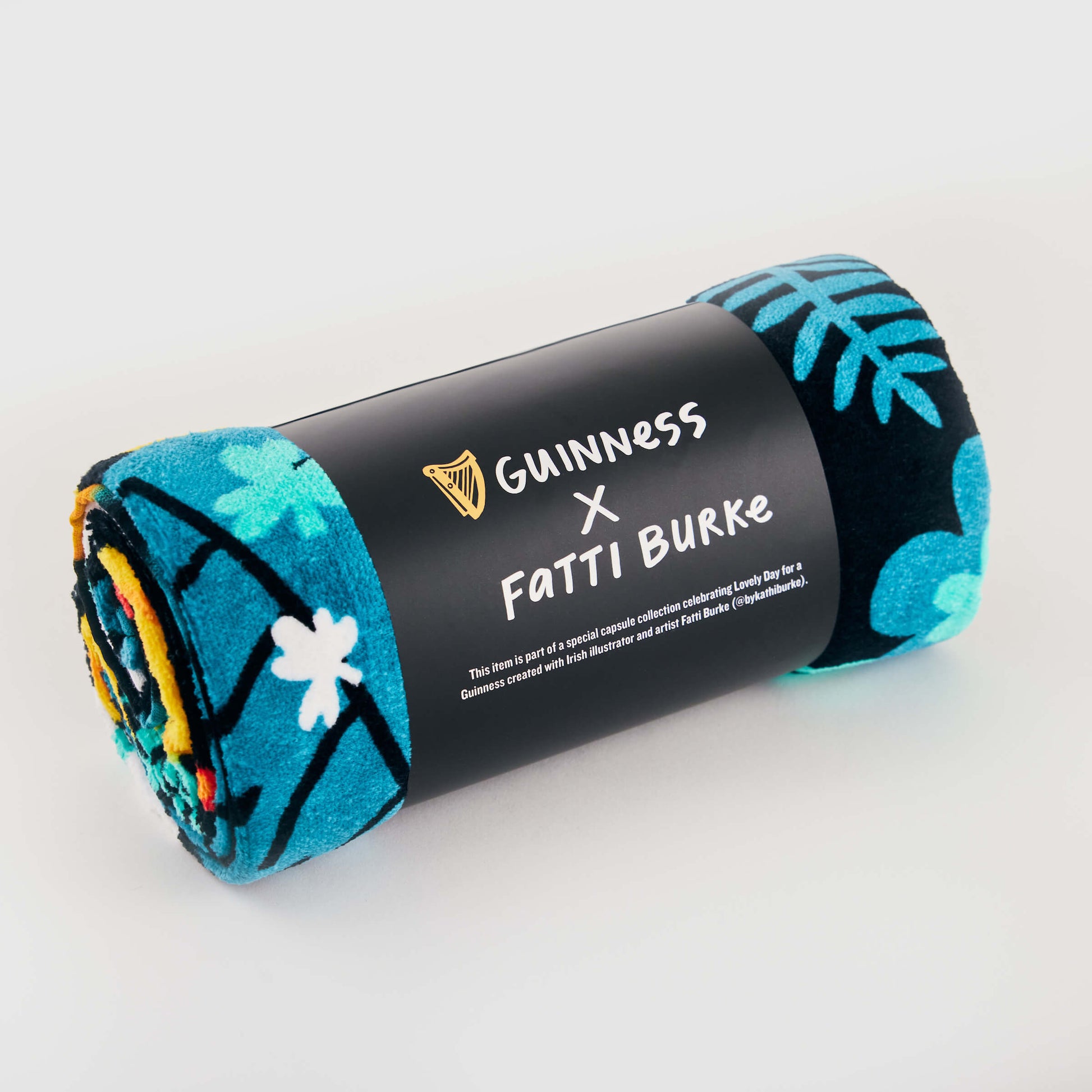 The Guinness x Fatti Burke Hawaiian Beach Towel features a blue and teal floral print, rolled and wrapped with a branded black band—ideal for stylish beach relaxation.