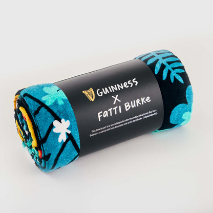The Guinness x Fatti Burke Hawaiian Beach Towel features a blue and teal floral print, rolled and wrapped with a branded black band—ideal for stylish beach relaxation.