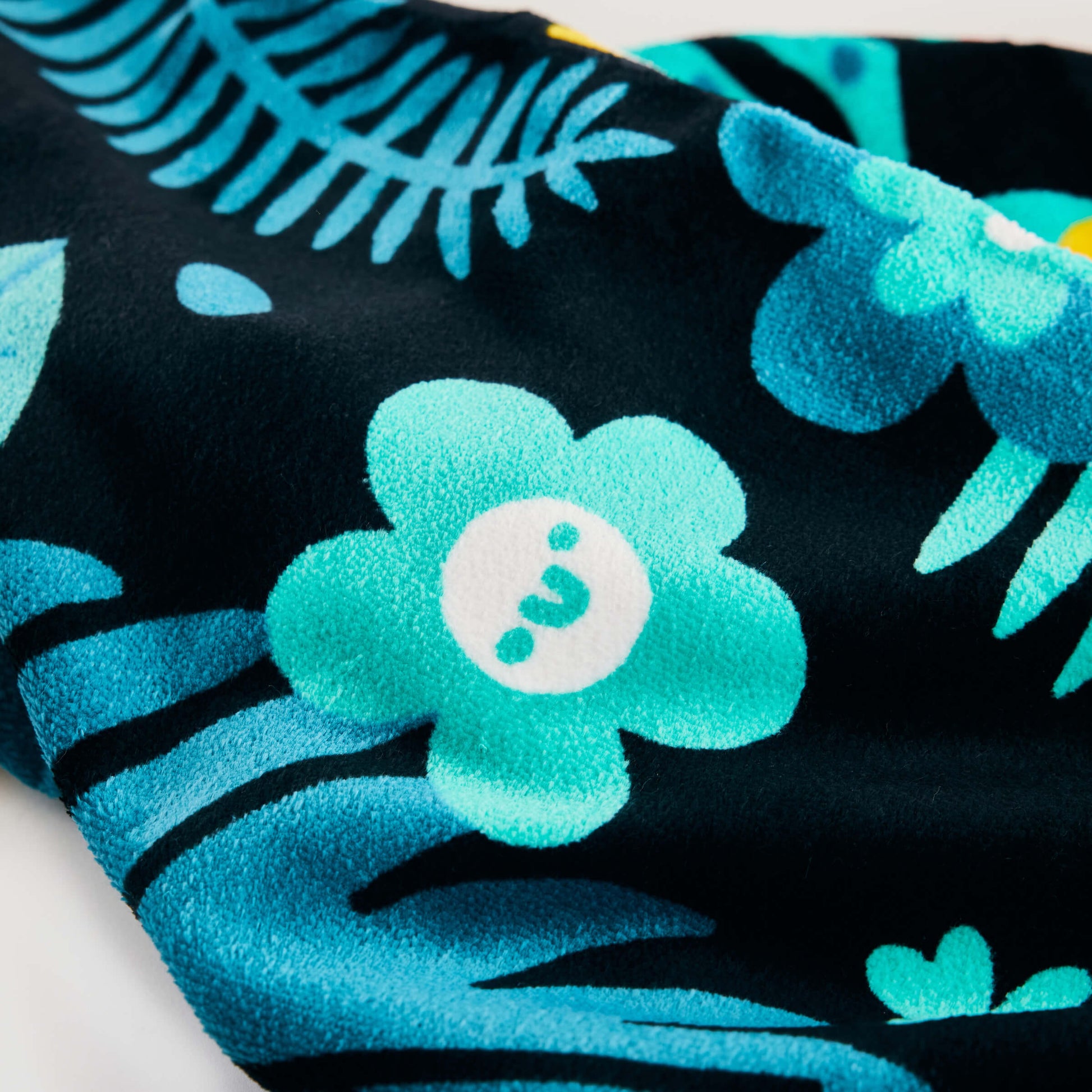 Close-up of the Guinness x Fatti Burke Hawaiian Beach Towel by Guinness, featuring a blue smiling flower and leaf pattern on a black background.