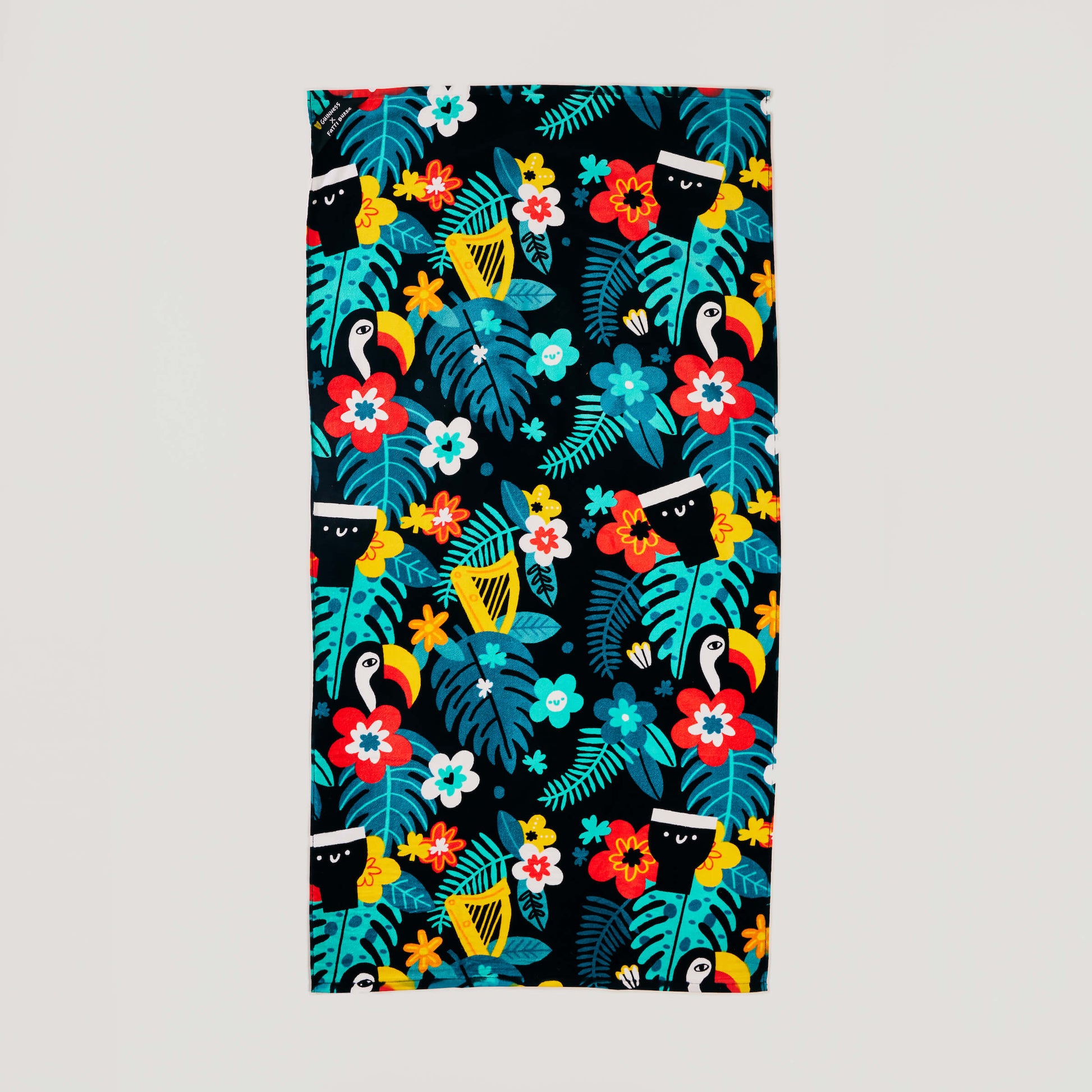 The Guinness x Fatti Burke Hawaiian Beach Towel is a rectangular cotton towel featuring a colorful Hawaiian pattern with leaves, flowers, toucans, and harps on a black background.