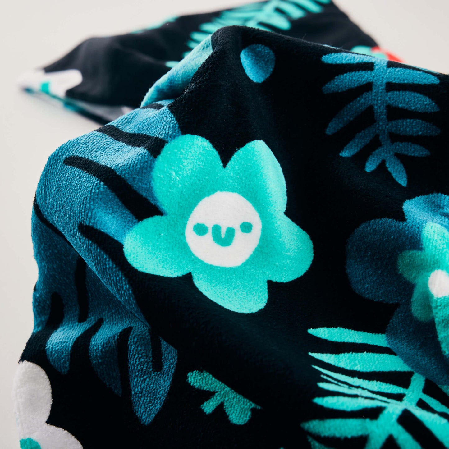 A close-up of the Guinness x Fatti Burke Hawaiian Beach Towel by Guinness, showing soft black cotton with a smiling blue flower, teal leaves, and a vibrant Hawaiian pattern.