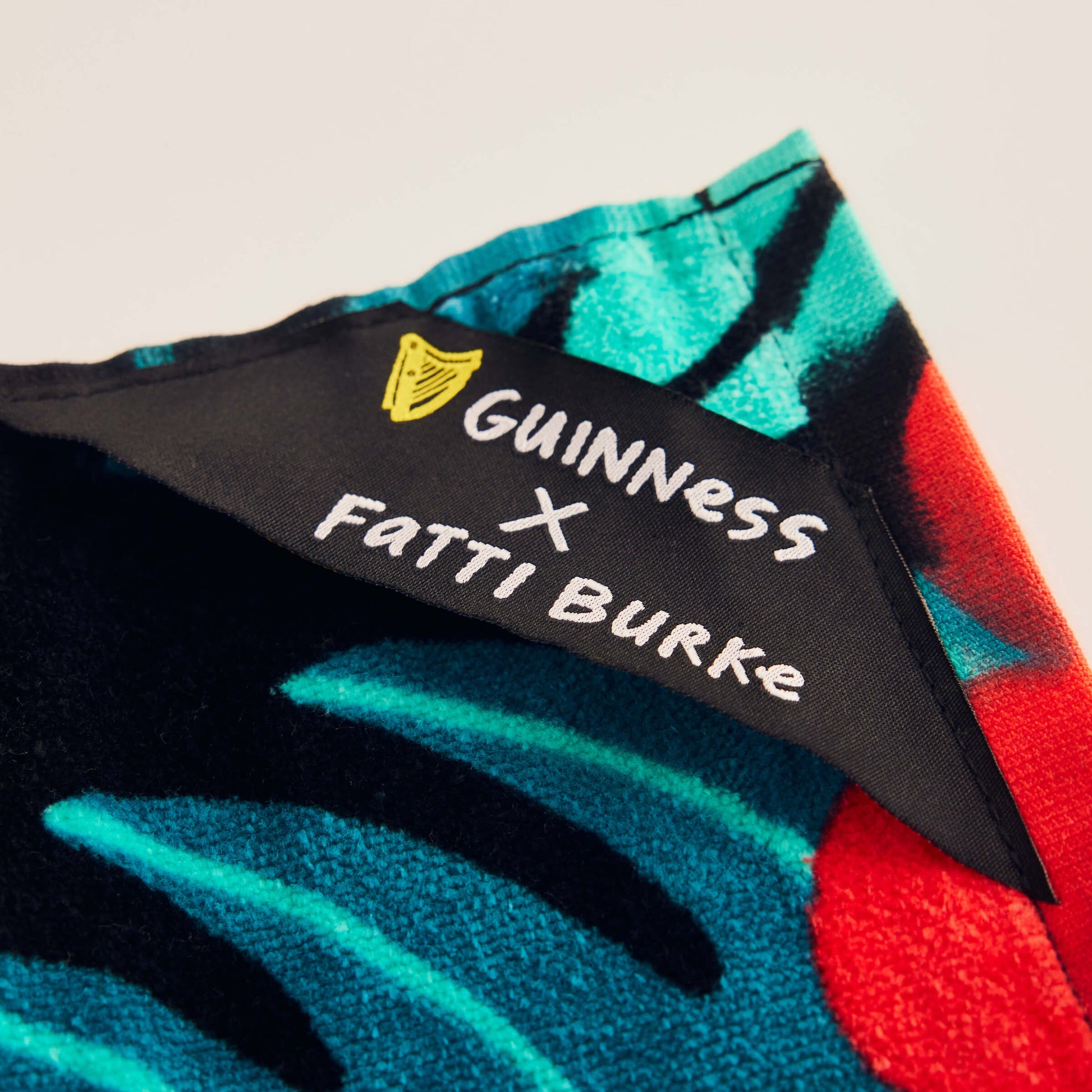 The Guinness x Fatti Burke Hawaiian Beach Towel features a colorful abstract pattern and a close-up of the corner shows a black label with a yellow harp and “GUINNESS x FATTI BURKE” text.