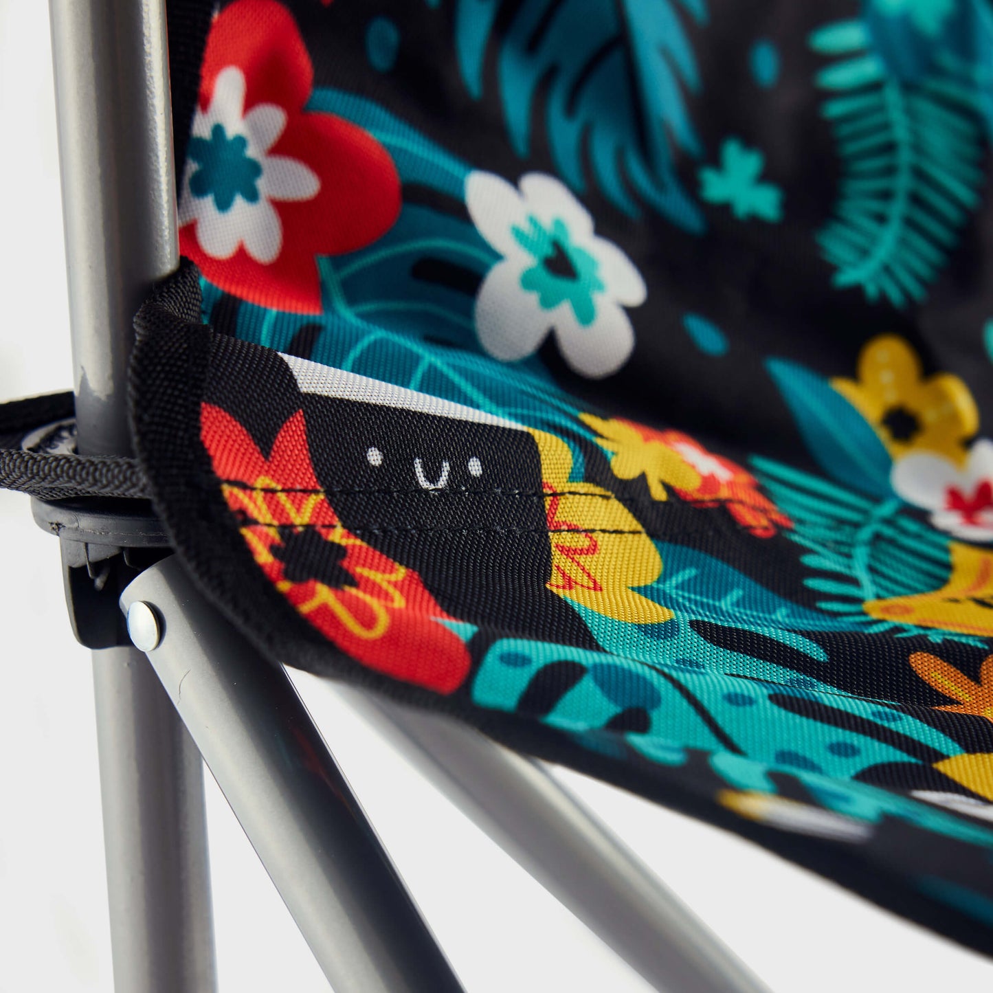 Close-up of the Guinness x Fatti Burke Hawaiian Camping Chair, featuring a black frame and vibrant floral fabric inspired by Hawaiian beach towels; a small tag with a smiling face is sewn into the seat. Brand: Guinness.