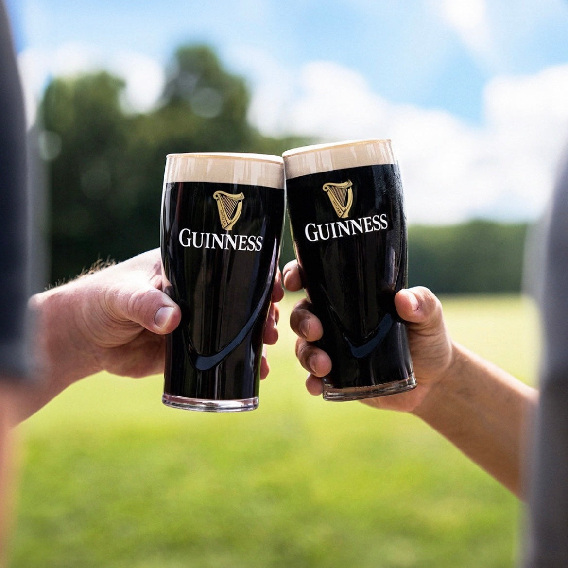 Two people clink glasses from the Guinness Gravity Pint Glass 2 Pack Gift Set by Guinness outdoors, with a green field and trees softly blurred in the background.
