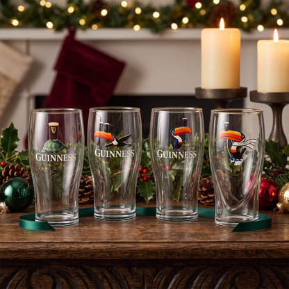 The Guinness Gilroy Christmas Glass Set from Guinness Webstore US is displayed on wood, a perfect festive gift among holiday décor and glowing candles.