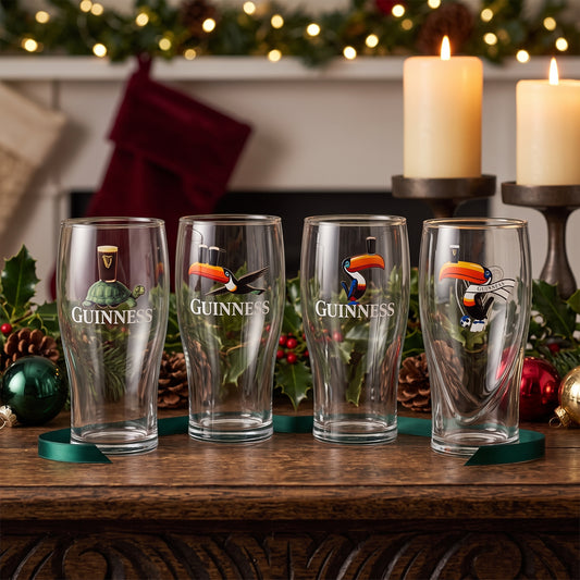 The Guinness Gilroy Christmas Glass Set from Guinness Webstore US is displayed on wood, a perfect festive gift among holiday décor and glowing candles.