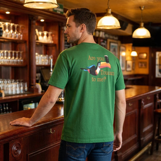 A man stands at a wooden bar wearing the Guinness Gilroy Toucan Tee – Green from Guinness Webstore US, featuring a toucan graphic and "Are you Toucan to me?" printed on the back.