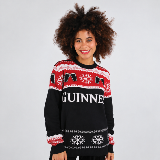 A woman with curly brown hair wearing a black, red, and white Guinness Christmas sweater, standing against a light gray studio background.