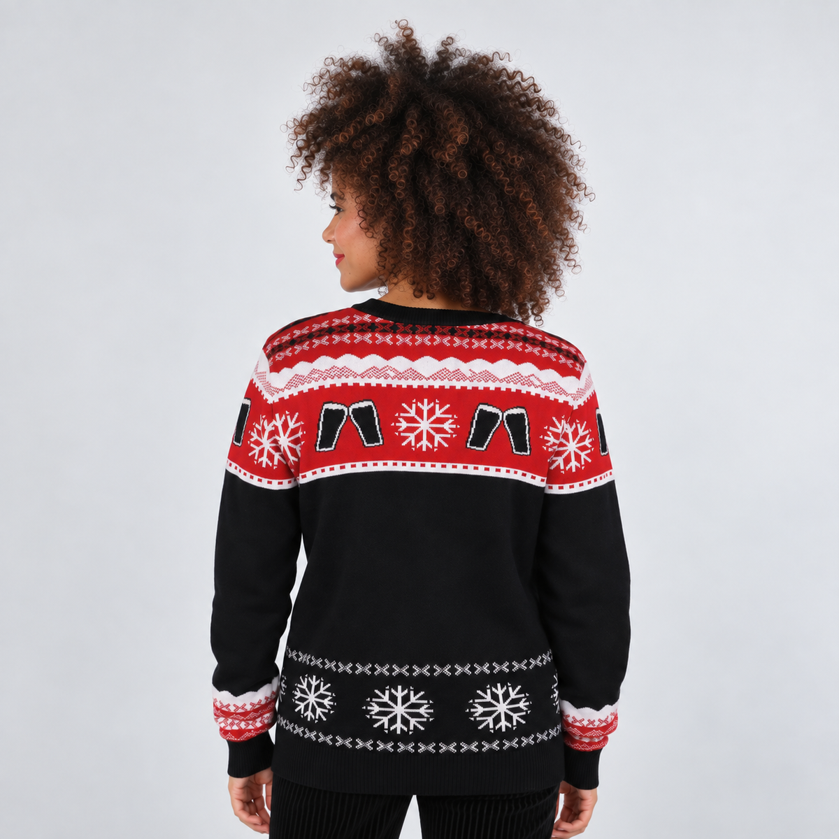 A woman viewed from the back wearing a black knit Christmas sweater with red and white Nordic-style patterns across the shoulders and upper back, against a light gray studio background.