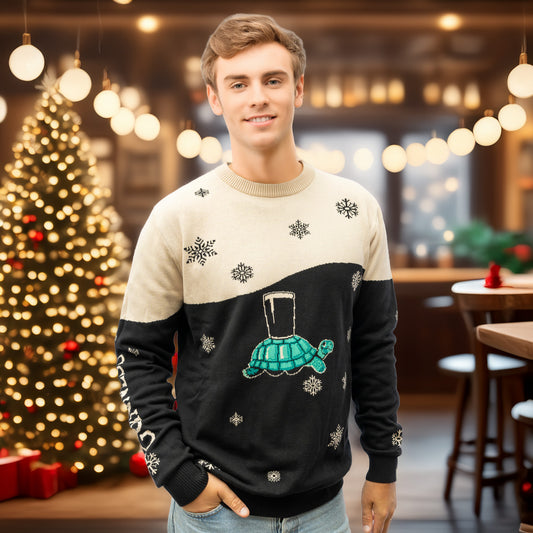 A young man stands indoors by a decorated Christmas tree, wearing the Guinness Holiday Black and Cream Tortoise Sweater from Guinness Webstore US, surrounded by festive lights and decor.