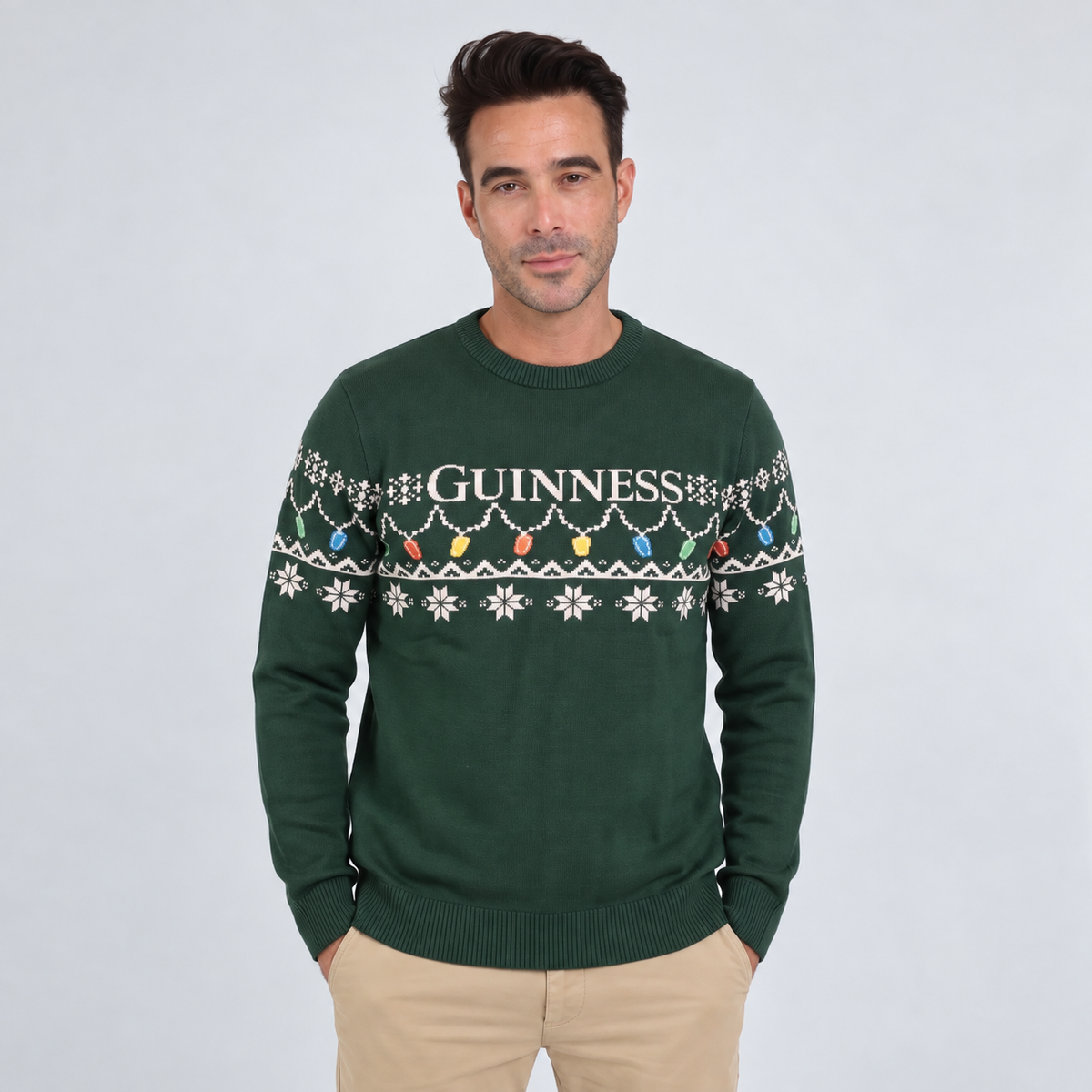 A man wearing the Guinness Holiday Pint Lights Sweater in green by Guinness Webstore US, which features “Guinness” text and a festive pattern, standing against a plain light background.
