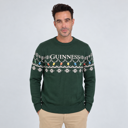 A man wearing the Guinness Holiday Pint Lights Sweater in green by Guinness Webstore US, which features “Guinness” text and a festive pattern, standing against a plain light background.