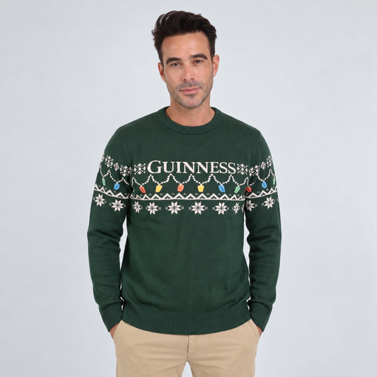 A man wearing the Guinness Holiday Pint Lights Sweater in green by Guinness Webstore US, which features “Guinness” text and a festive pattern, standing against a plain light background.