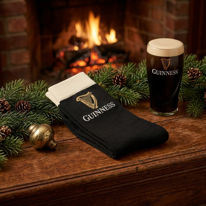 Guinness Pint Crew Socks – Black with a cream-colored cuff and gold harp logo, displayed on a wooden mantle next to a pint of stout, pine branches, and a fireplace.