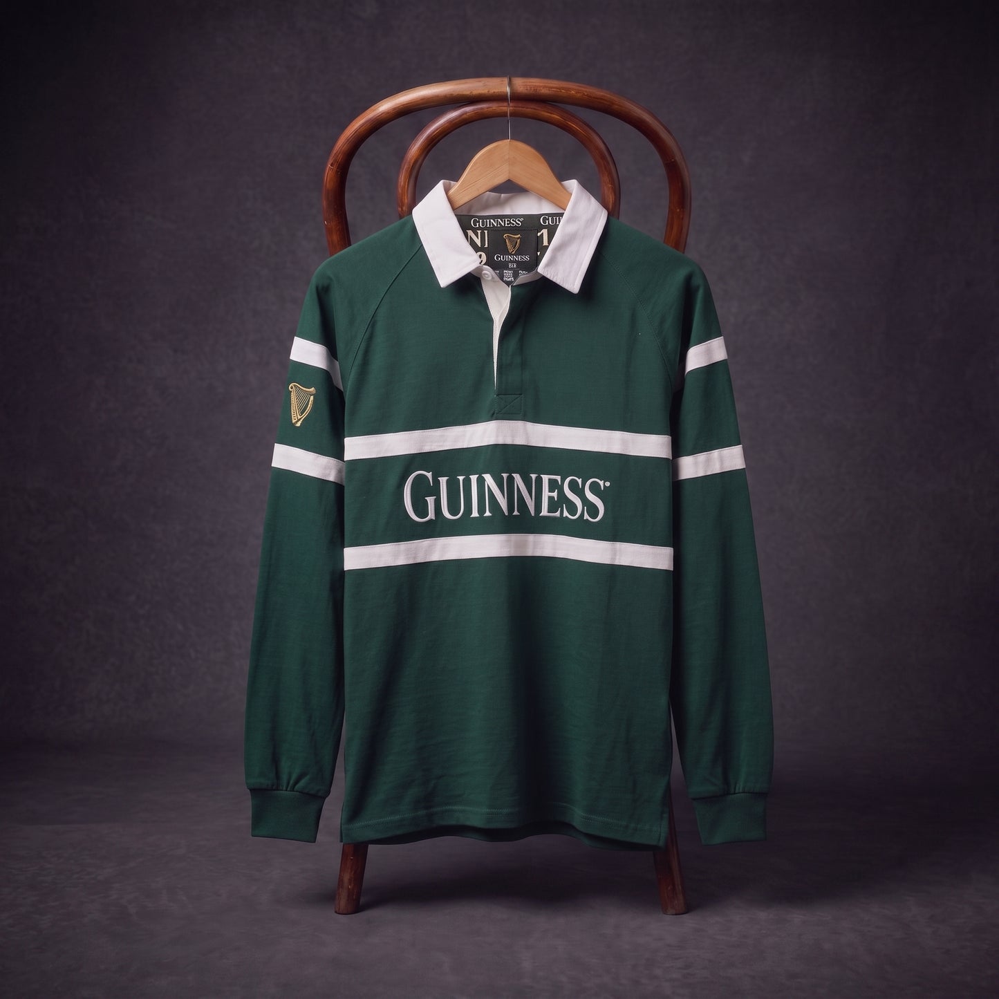 A Guinness Green Traditional Rugby Jersey with white stripes and “Guinness” printed on the front hangs on a wooden chair against a dark background.