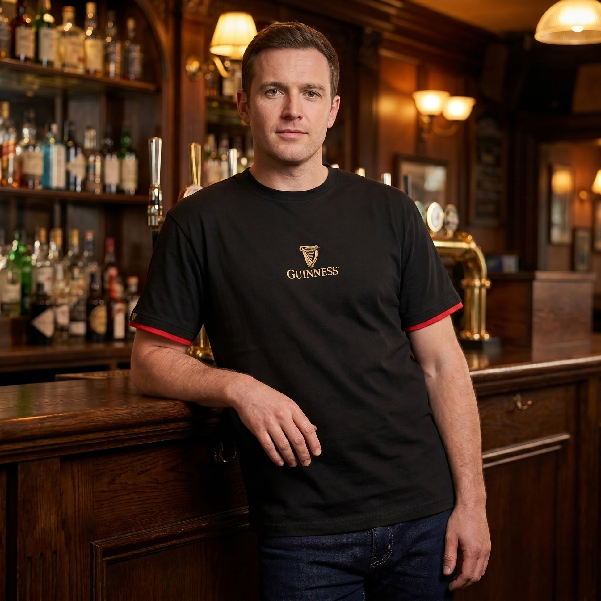 A man wearing the Guinness Gaelic Label Black Tee from Guinness Webstore US stands by a wooden bar in a pub, with shelves of bottles and beer taps in the background.