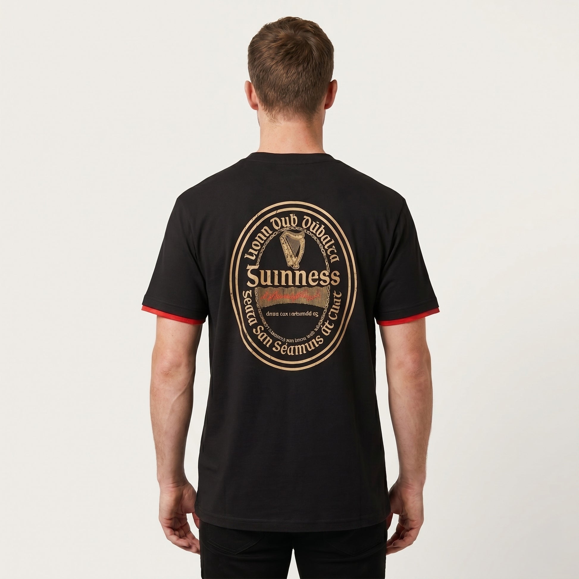 A person stands facing away, wearing the Guinness Gaelic Label Black Tee from Guinness Webstore US, featuring a prominent gold and red Guinness logo and text on the back.