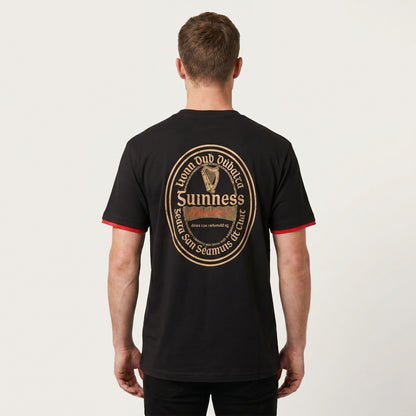 A person stands facing away, wearing the Guinness Gaelic Label Black Tee from Guinness Webstore US, featuring a prominent gold and red Guinness logo and text on the back.