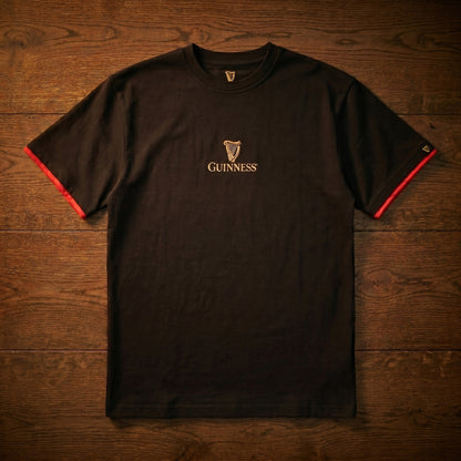 The Guinness Gaelic Label Black Tee from Guinness Webstore US features the iconic logo and harp on the chest, red sleeve trim, and a classic Irish heritage style—shown laid flat on a wooden surface.