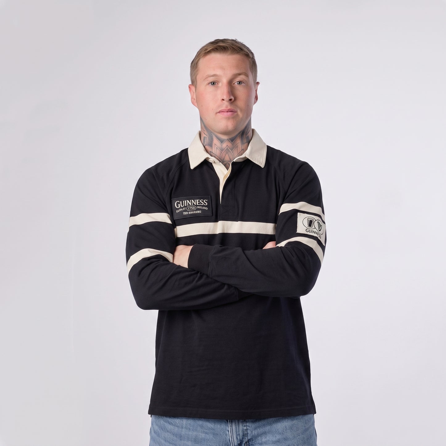 A man with short hair and visible neck tattoos stands with his arms crossed, wearing the Guinness Black Traditional Rugby Jersey by Guinness—crafted from 100% cotton—in black and white, paired with blue jeans against a plain white background.