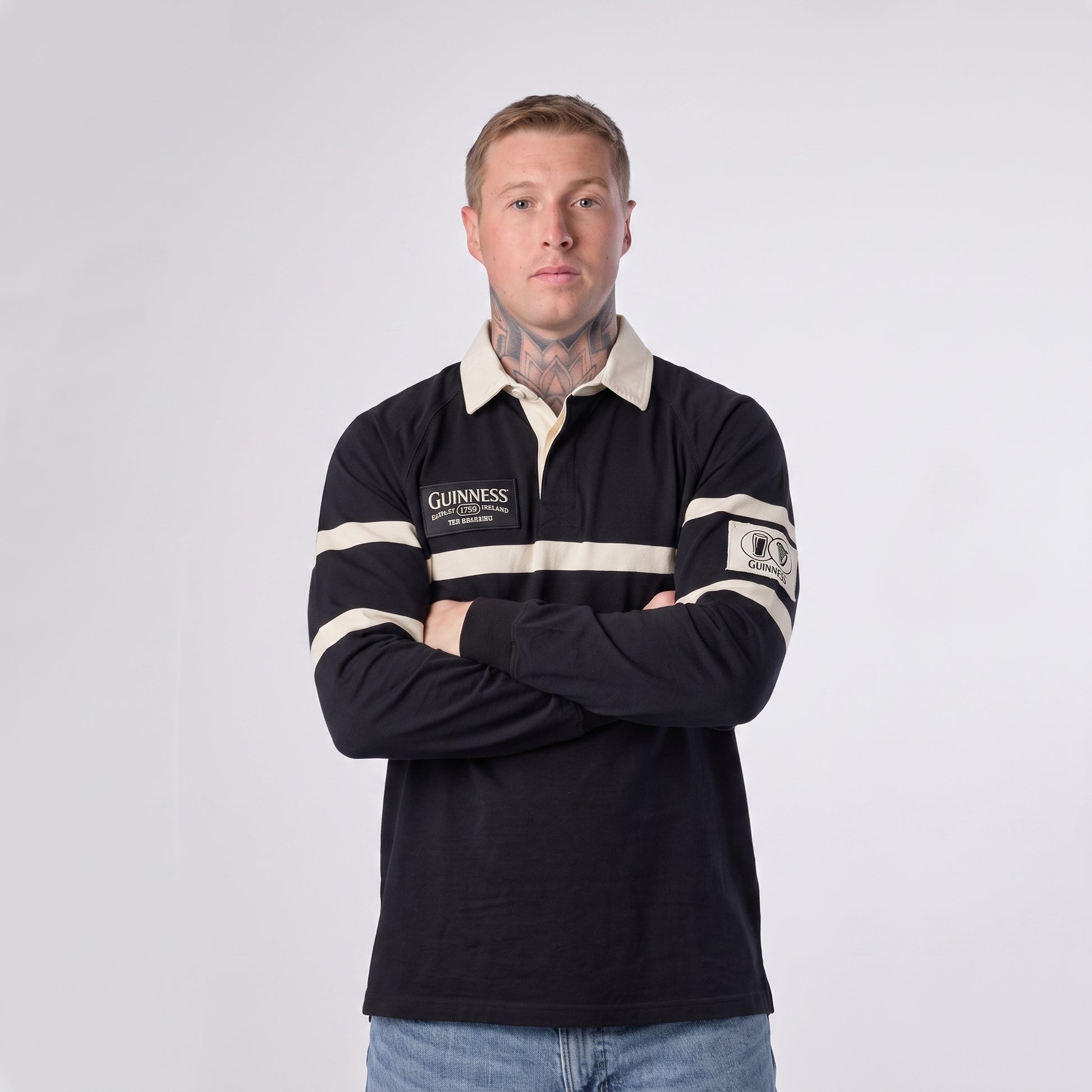 A man with short hair and visible neck tattoos stands with his arms crossed, wearing the Guinness Black Traditional Rugby Jersey by Guinness—crafted from 100% cotton—in black and white, paired with blue jeans against a plain white background.