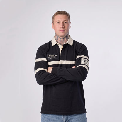 A man with short hair and visible neck tattoos stands with his arms crossed, wearing the Guinness Black Traditional Rugby Jersey by Guinness—crafted from 100% cotton—in black and white, paired with blue jeans against a plain white background.