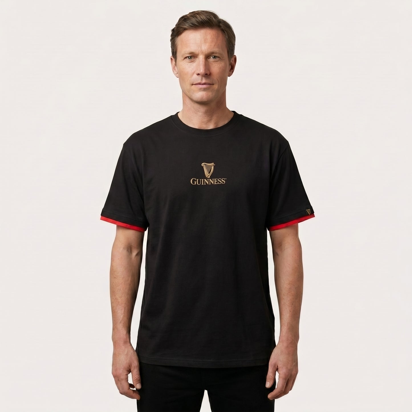 A man faces forward against a plain light background, wearing the Guinness Gaelic Label Black Tee from Guinness Webstore US, featuring red sleeve accents.