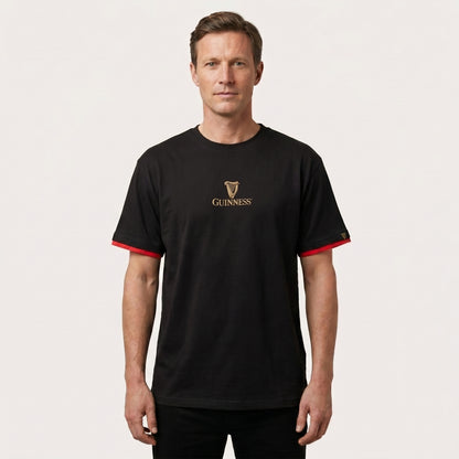 A man faces forward against a plain light background, wearing the Guinness Gaelic Label Black Tee from Guinness Webstore US, featuring red sleeve accents.